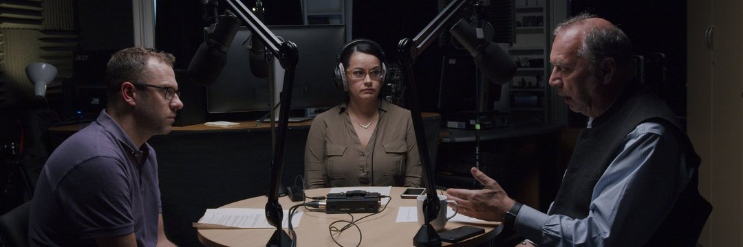 Image of three people sitting at a round table with microphones. The man on the left in a purple shirt is looking across the table to a man gesturing with his hands and speaking, while a woman in a green shirt looks on.