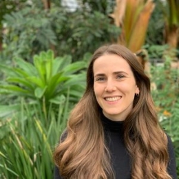 White woman with long brown hair in front of green foliage