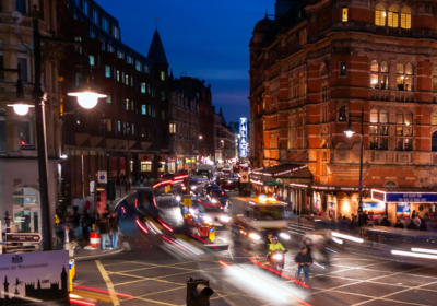 Pedestrians wait at a crossing as traffic drives past at night. 